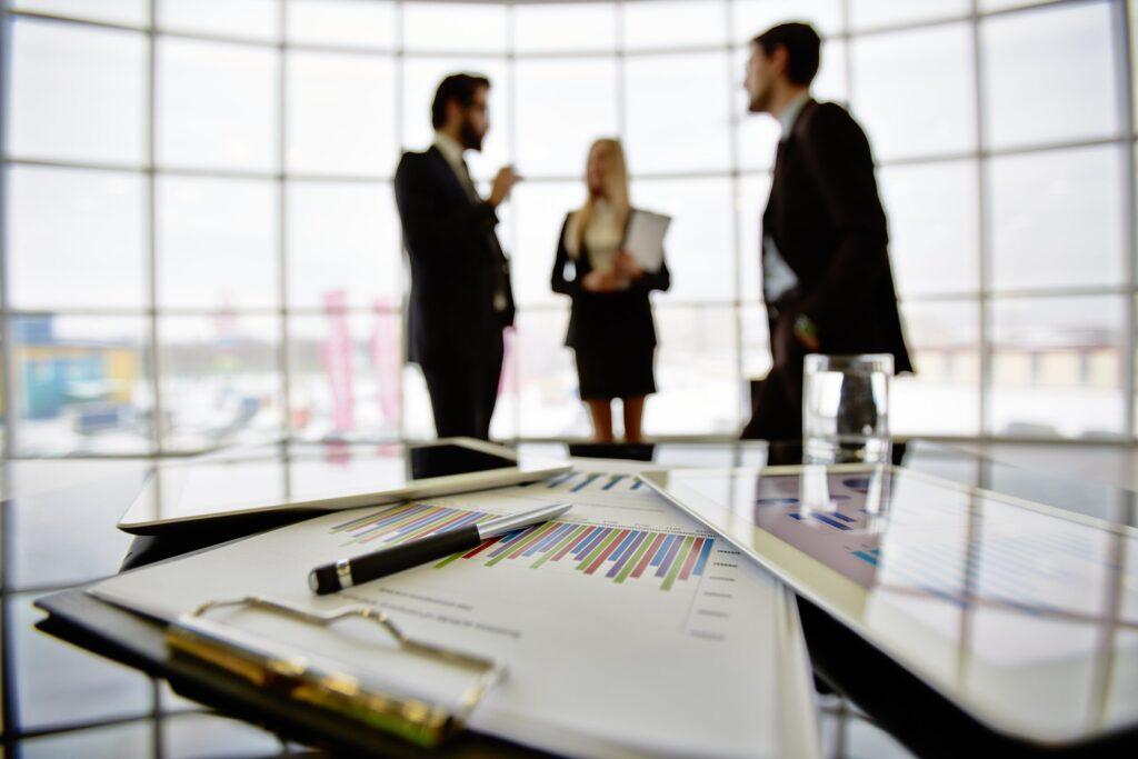 Business documents with bar charts, tablet, and pen on a table, with professionals discussing in the background, symbolizing transfer pricing advantages