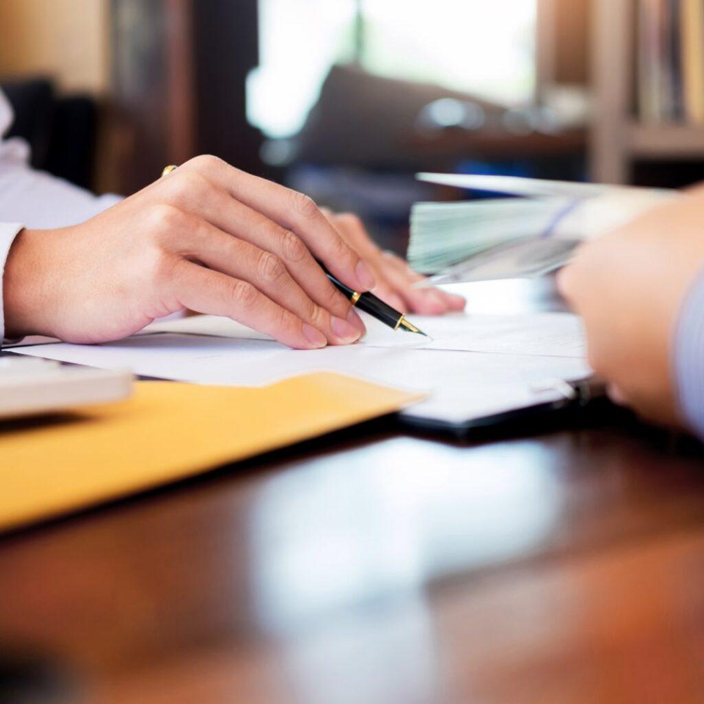 Close-up of businessperson reviewing documents with a pen and exchanging a stack of money, symbolizing transfer pricing trends and financial transactions.