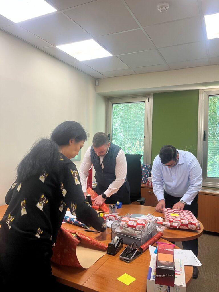 Office colleagues wrapping Christmas gifts at a desk, using festive paper and ribbons.