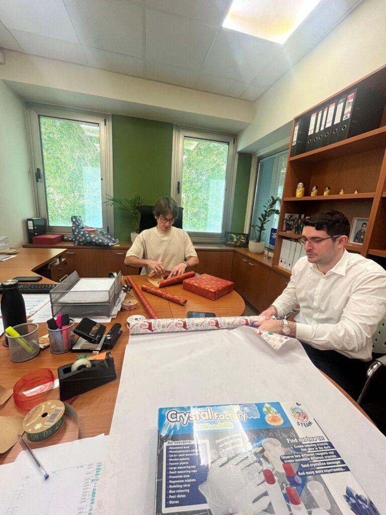 Office colleagues wrapping Christmas gifts together at a desk, preparing for a festive celebration.